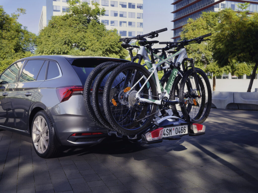 Naložená elektrokola na nosiči tažného zařízení u šedého auta Škoda před cestou.