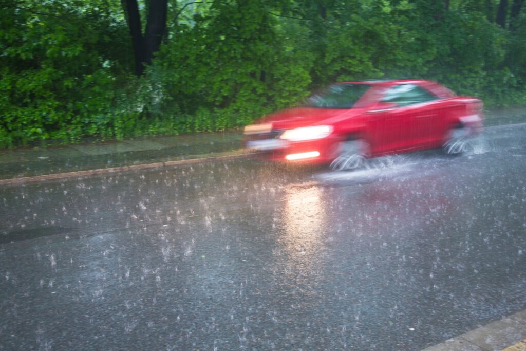 Červené auto jedoucí za silného deště na mokré dálnici, se zapnutými světly a viditelným rozstřikem vody od kol, což ukazuje reálné riziko aquaplaningu v provozu.