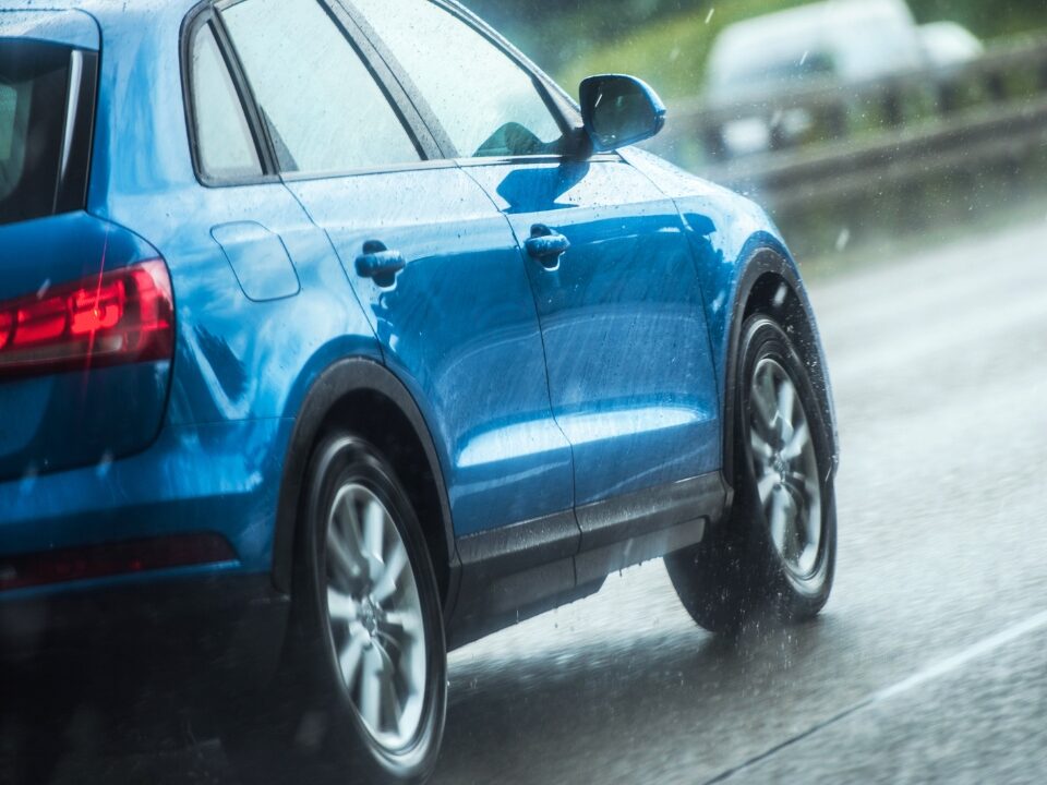 Akční pohled na zadní část jedoucího modrého SUV, jak jeho pneumatiky za deště odhazují vodu, což ilustruje okamžik před ztrátou přilnavosti a vznikem aquaplaningu.